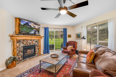 A cozy living room with a ceiling fan, stone fireplace, large window, leather sofa, armchairs, wooden coffee table, and TV mounted above the fireplace. Blue curtains frame a view into a green yard.