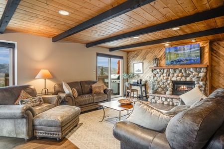 A cozy living room with wooden ceiling beams, a stone fireplace, and leather sofas. A framed picture hangs above the fireplace, and a sliding glass door provides access to an outdoor patio.