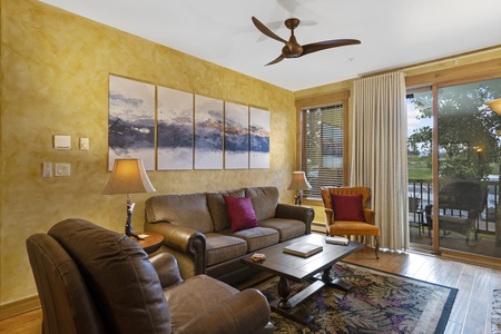 Cozy living room with brown leather sofas, wooden coffee table, colorful rug, and large windows with sheer curtains. A ceiling fan and triptych artwork adorn the space.