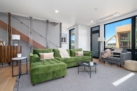 A modern living room with a green sectional sofa, a gray armchair, and a round rug. Large windows let in natural light and provide an exterior view. A wooden staircase with a metal mesh railing is visible.