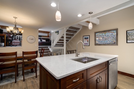 Modern kitchen with wooden cabinets, a marble countertop island with a built-in sink, and a dishwasher. Adjacent dining area with a wooden table and chairs, and stairs leading to an upper level.