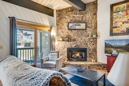 A cozy living room with a stone fireplace, comfortable furniture, a Colorado sign above the mantel, and a mountain-themed wall art. A sliding glass door opens to a balcony with a view.