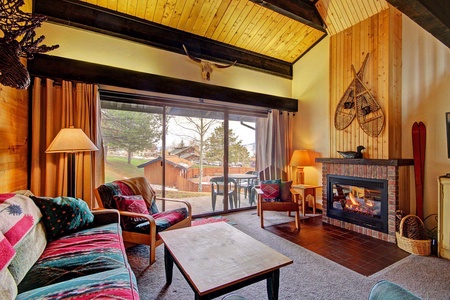 A cozy living room with a vaulted wooden ceiling, a fireplace, colorful furniture, and large windows revealing a patio and outdoor scenery. Vintage snowshoes and antlers decorate the wall above the fireplace.