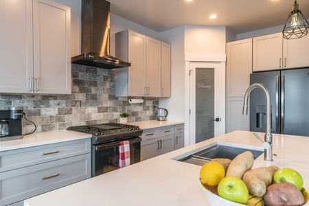 Modern kitchen with grey cabinets, stainless steel appliances, and a central island with a sink. A bowl of fruits and vegetables sits on the island. The backsplash features grey tiles, and the pantry door is frosted.