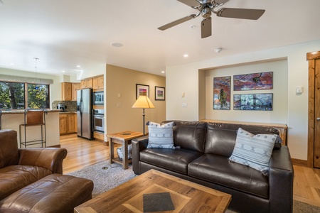 A modern living room with a brown leather couch, two armchairs, a wooden coffee table, and a kitchen in the background. Artworks hang on the wall above the couch. A ceiling fan and lamp are present.