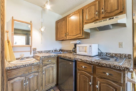 A compact kitchen with wooden cabinets, granite countertops, a small sink, a microwave, an electric stovetop, a mini fridge, a mirror on the wall, and a white light fixture.