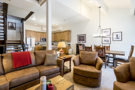 A cozy living room with brown sofas and chairs, a wooden coffee table, framed artwork, a staircase, a dining area with a chandelier, and a kitchen in the background with wooden cabinetry.