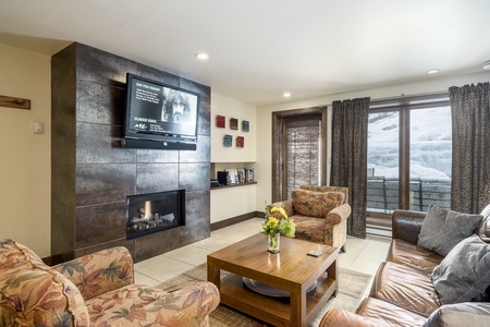 A cozy living room featuring a wall-mounted TV above a fireplace, two patterned armchairs, a sofa, a wooden coffee table with a flower vase, and large windows with a snowy view outside.