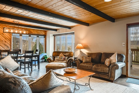 A cozy living room with brown leather sofas, a wooden coffee table, a table lamp, and a dining area in the background, featuring large sliding glass doors overlooking an outdoor deck.