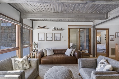 A cozy living room with large windows, a brown leather sofa, two gray armchairs with patterned pillows, and a round ottoman. Walls are decorated with framed pictures and shelves. Bedroom visible through the door.