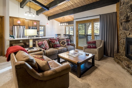 A cozy living room with wooden ceiling, exposed beams, and carpeted floor. It features a stone fireplace, brown leather sofas with red pillows, a wooden coffee table, and a kitchen in the background.