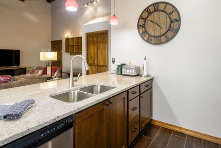 A kitchen with a large countertop featuring a double sink, dark wooden cabinets, and a dishwasher. In the background are a living area, a large wall clock, and pendant lights hanging from the ceiling.