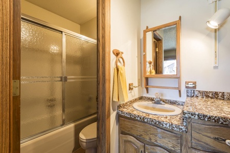 An interior bathroom features a glass-enclosed bathtub with a shower, a toilet, a granite countertop with a built-in sink, a wooden vanity, and a wall-mounted mirror. Yellow towels hang by the sink.