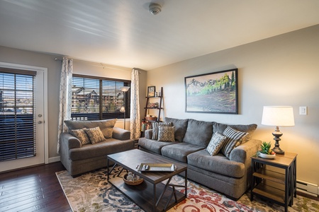 A cozy living room with a gray sofa and loveseat, a coffee table, a rug, a framed mountain painting, a lamp on a side table, and windows with blinds and drapes.