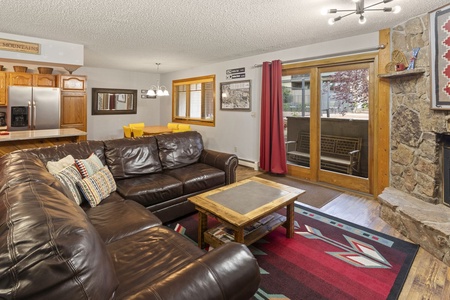 A living room with a brown leather sectional sofa, stone fireplace, wooden coffee table, and sliding glass door opening to a balcony. The dining area and kitchen are visible in the background.