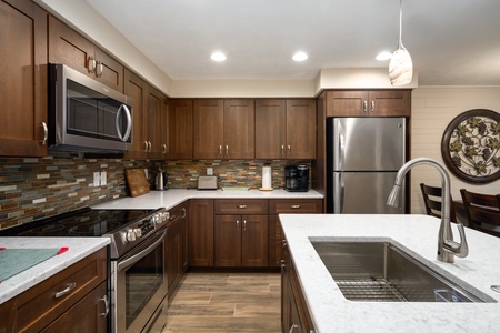 Modern kitchen with dark wooden cabinets, stainless steel appliances including a refrigerator and microwave, a sink in a central island, and a mosaic tile backsplash under bright ceiling lights.
