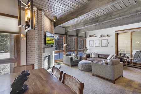 A cozy living room with a modern design, featuring wooden ceiling beams, a brick fireplace, a wall-mounted TV, large windows, a leather sofa, two armchairs, and a dining table with chairs.