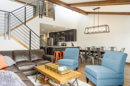 A modern living room with brown leather sofa, two blue chairs, a wooden coffee table, open kitchen, dining area with metal chairs, and staircase leading to an upper level.