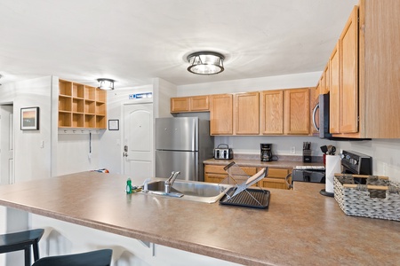 Modern kitchen with wooden cabinets, stainless steel appliances, and a central island featuring a sink and dish rack. Shelving and coat hooks are mounted on the wall near the entrance.