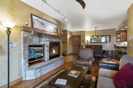 A cozy living room with a stone fireplace, a television, a plush sofa, and various decorative elements. The room features warm lighting and natural wood accents throughout.