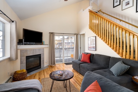 A cozy living room features a grey sectional sofa with red and polka dot pillows, a fireplace, a wall-mounted TV, and a coffee table. Upstairs, a wooden staircase is visible. Large sliding doors let in natural light.