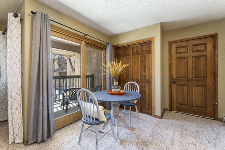 A small dining area with a round table and two chairs in front of sliding glass doors leading to a balcony. A decorative vase with dried wheat and a bowl of oranges are on the table.