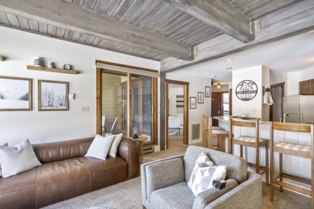 A cozy living area with a brown leather couch, a gray armchair, a breakfast bar with three stools, and a kitchen in the background. The room features wooden beams and large framed photos on the walls.