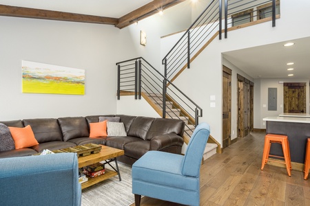 A modern living room with a brown leather sectional sofa, blue chairs, wood coffee table, and a staircase with black railings leading to the upper floor. The space is brightly lit with recessed lighting.