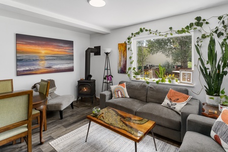 A cozy living room with a gray sofa, a wooden coffee table, a small dining area, plants on the windowsill, wall art, and a wood-burning stove. A window reveals an outdoor view.