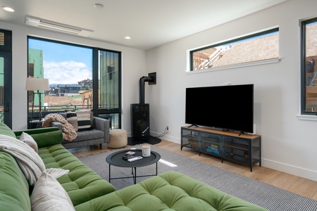 A modern living room features a green sofa, a gray armchair, a black round coffee table, a flat-screen TV, large windows, and wood flooring.