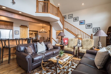 A spacious living room features leather sofas, a wooden staircase, artwork on the walls, and a kitchen with bar seating in the background. Natural light brightens the space.