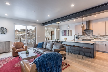 A modern open-concept living room and kitchen featuring a brown leather sofa, red area rug, a central coffee table, bar stools at the kitchen island, and large windows with a snowy outdoor view.