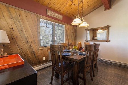 A dining area with a wooden table set for five, chairs, a lamp, windows with blinds, and a wall mirror reflecting part of the room. The ceiling is wood-paneled with two hanging lights. A sign reads "Mountain Rules.