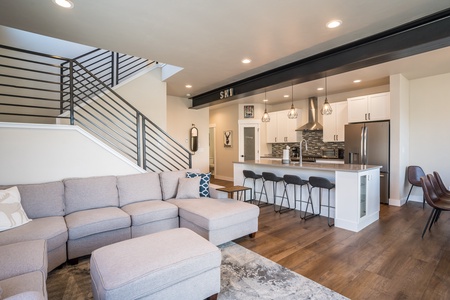 Modern open-concept living area with a gray sectional sofa, a kitchen with a breakfast bar and stools, and a staircase with a metal railing.