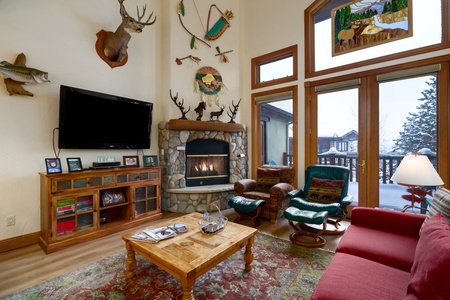 A cozy living room with a stone fireplace, mounted animal heads, and large windows. There is a red sofa, two green chairs, a wooden coffee table, and a TV on a wooden media console.