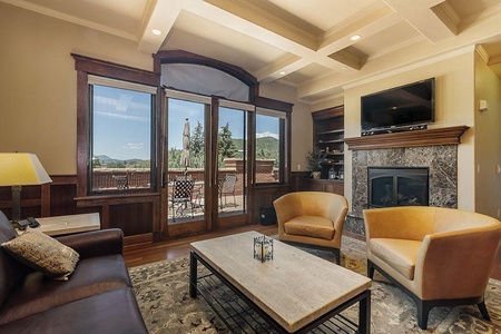 A cozy living room features a brown leather couch, two tan chairs, a marble coffee table, a fireplace, a wall-mounted TV, and large windows with a view of a deck and mountains.