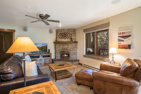 A cozy living room with a leather sofa, armchair, wooden coffee tables, a stone fireplace, a TV on a wooden stand, and a window with outdoor views. Ceiling fan and lamps provide lighting.