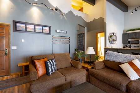 A living room with brown sofas, a chalkboard, and a framed picture on a wall with a mountain mural. The space includes a kitchen with dark cabinets and a small bar area.