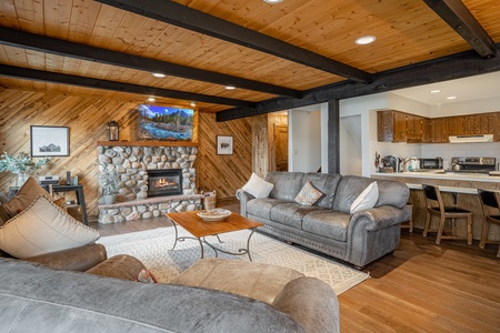 A cozy living room with wooden walls, a stone fireplace, two gray sofas, a wooden coffee table, and a TV mounted above the fireplace. The room opens into a kitchen with wooden cabinets.