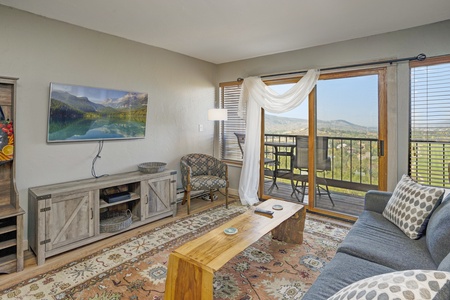 A cozy living room with a flat-screen TV, a wooden coffee table, a patterned rug, a sofa, an armchair, and sliding doors leading to a balcony with a scenic view.