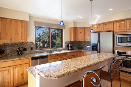 A modern kitchen with wooden cabinets, granite countertops, stainless steel appliances, and a large island with seating. The room is well-lit with natural light from a window and pendant lights.