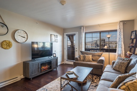 A cozy living room with a light gray sofa, matching armchair, wooden coffee table, flat-screen TV on a gray stand with a built-in fireplace, wall clock, decorative items, and large window with curtains.