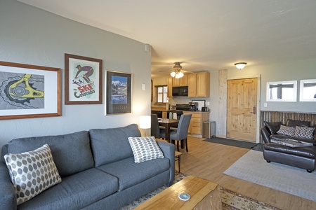 Living room with a gray couch, patterned pillows, wall art, and a small wooden coffee table. The room leads into an open kitchen area with wooden cabinets and a dining table.