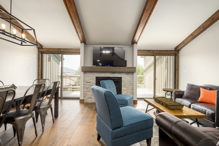 A living room with a fireplace under a wall-mounted TV, a dining area with metal chairs, blue armchairs, a brown leather couch, and sliding doors opening to a wooden deck.