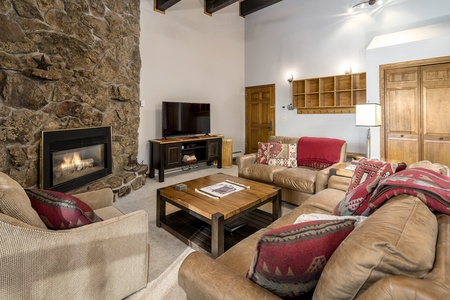 A cozy living room with a stone fireplace, beige sofas adorned with red and beige patterned cushions, a wooden coffee table, and a TV on a black stand. Shelves and closed doors are in the background.