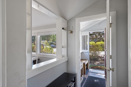 View of a small entryway with an open door leading outside. The space features a coat hook, bench, and large windows. Light pours in from the outside, creating a bright and welcoming atmosphere.