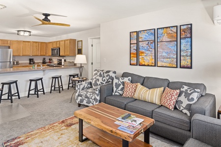 A modern living room with a gray sofa, patterned chairs, a wooden coffee table, and a kitchen in the background. Art pieces are hung on the wall above the sofa, and a ceiling fan is installed.