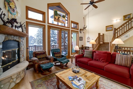 A cozy living room with large windows, a red sofa, two armchairs, a stone fireplace, and a wooden coffee table. A staircase leads up to a loft area, and multiple artworks decorate the walls.