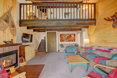 Cozy living room with wood accents, a fireplace, a mounted TV, a staircase leading to a loft, and rustic furniture including two couches, an armchair, and a wooden coffee table.