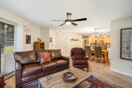 A living room with a brown leather sofa, red armchair, wooden coffee table, and ceiling fan. The room leads into a dining area with a table and chairs and a kitchen with wooden cabinets.